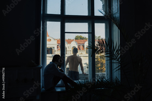 Romantic young couple relax at home. Man is standing on the on the balcony with cityscape and woman sitting on the windowsill.