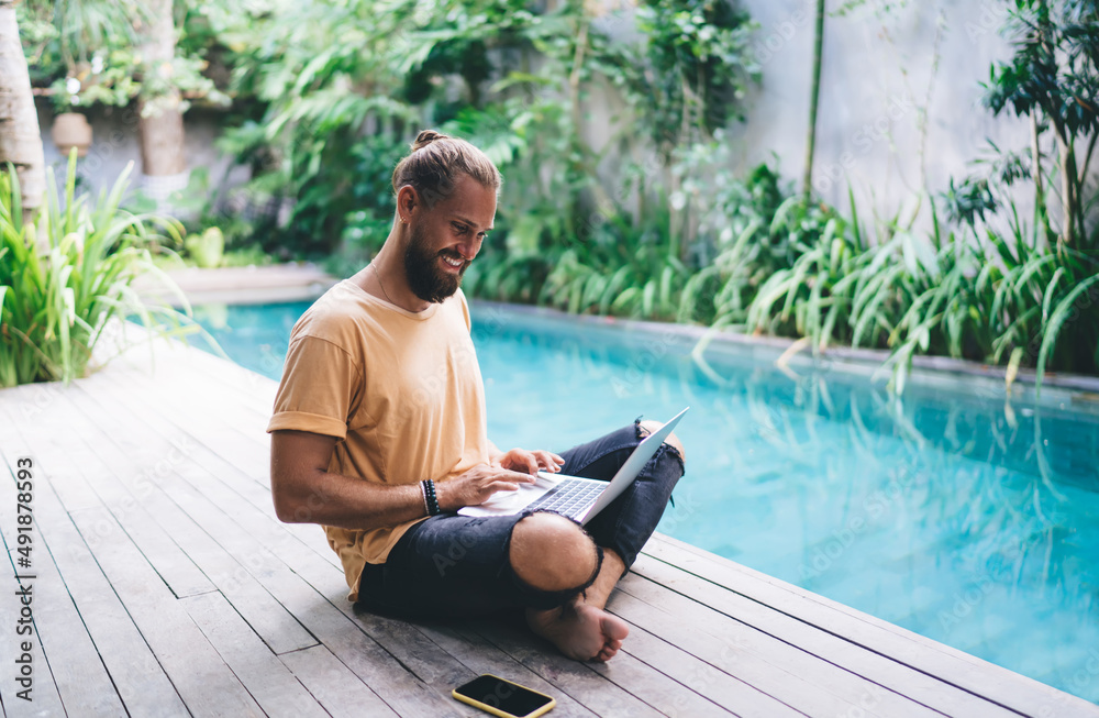 Cheerful male freelancer browsing video content while networking in social media using netbook technology at pool terrace, happy hipster guy blogging online via laptop enjoying digital nomad lifestyle Cheerful male freelancer browsing video content while networking in social media using netbook technology at pool terrace, happy hipster guy blogging online via laptop enjoying digital nomad lifestyle