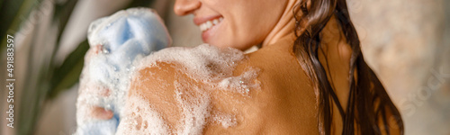 Happy young woman applying shower gel on her body using loofah sponge while taking shower. Spa and body care concept photo