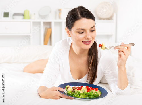 Pretty woman in white dress holding fork and eating vegetable salad in bedroom