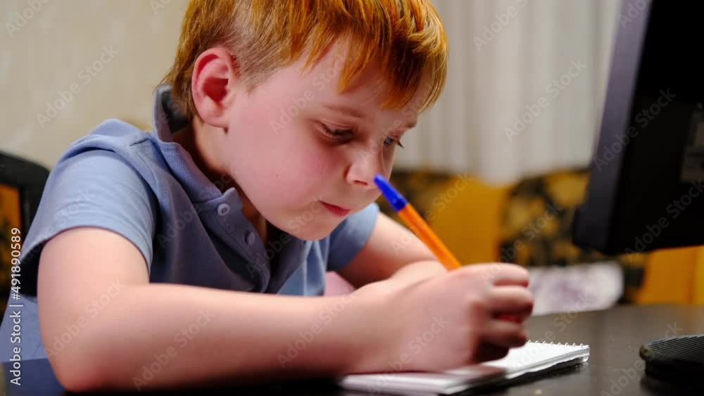 A young boy does homework, studies remotely, writes in a notebook in front of a computer screen. Home schooling. Quarantine. A young boy does homework, studies remotely, writes in a notebook in front of a computer screen. Home schooling. Quarantine.