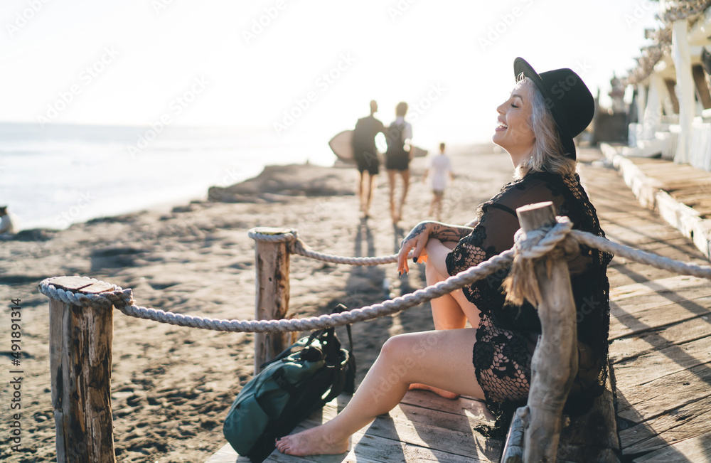 Fototapeta premium Happy cheerful model in stylish hat smiling during leisure time at sunny beach, joyful female dressed in bikini swimwear enjoying tropical vacations for recreation and sunbathing in Indonesia