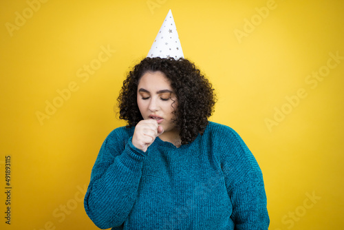 Young beautiful woman wearing a birthday hat over isolated yellow background with her hand to her mouth because she's coughing © Irene