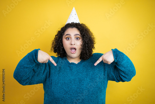 Young beautiful woman wearing a birthday hat over isolated yellow background looking confident with smile on face, pointing oneself with fingers proud and happy. © Irene