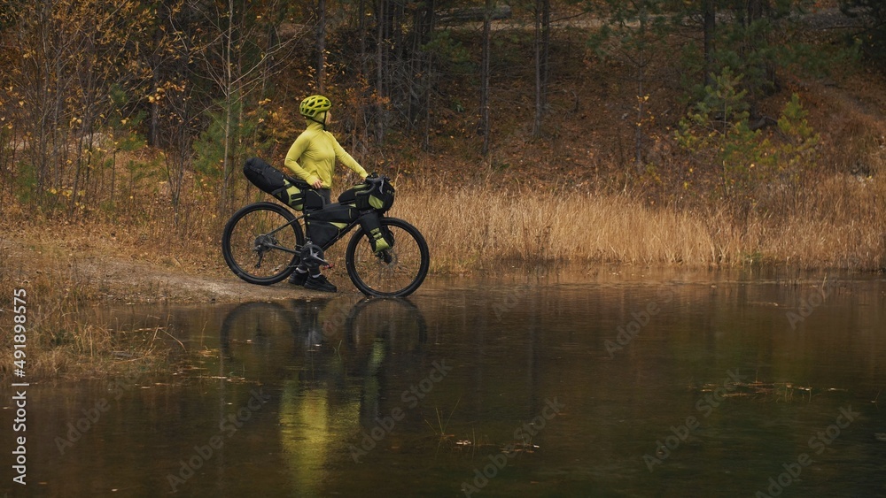 The woman travel on mixed terrain cycle touring with bikepacking. The traveler journey with bicycle bags. Sportswear in green black colors. The trip in magical autumn forest, river stream.