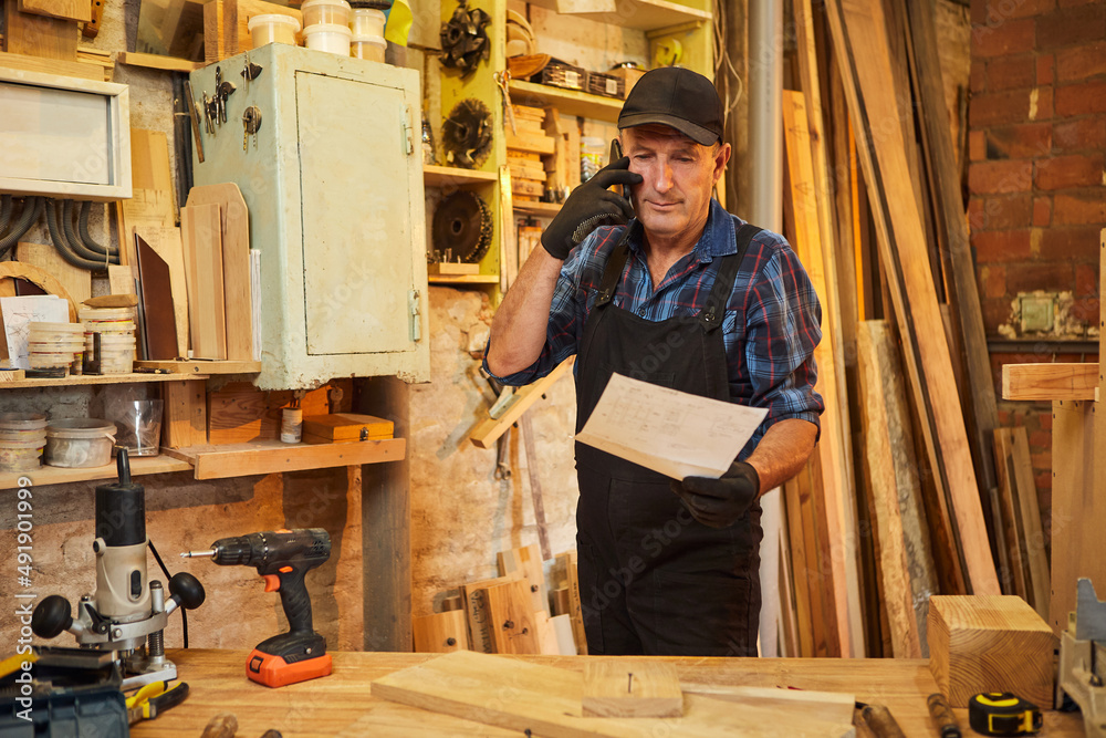 Portrait of a senior carpenter looking at blueprints plans to make a piece of furniture and talking on the phone with a client in the carpentry workshop Portrait of a senior carpenter looking at blueprints plans to make a piece of furniture and talking on the phone with a client in the carpentry workshop