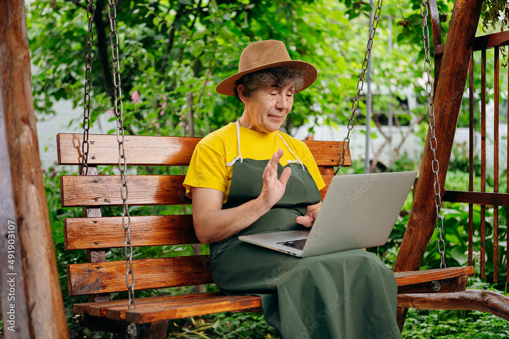 Senior woman gardener in a hat is watching something on a laptop computer and making a video call in the yard while outdoors.