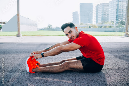 Portrait of strength Caucasian fitness trainer stretching legs and looking at camera during morning workout at urbanity, young muscular athlete in comfortable sportswear practice body flexibility