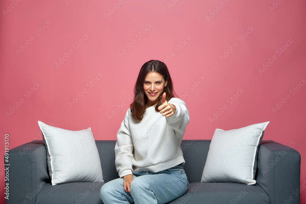 Confident satisfied young girl show thumb up, ask to put likes, advertise service, giving good feedback, sitting on sofa Confident satisfied young girl show thumb up, ask to put likes, advertise service, giving good feedback, sitting on sofa