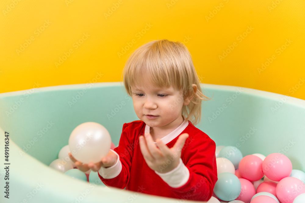 A little beautiful girl stands alone in dry pool with colorful balls and holds a white ball in her hands