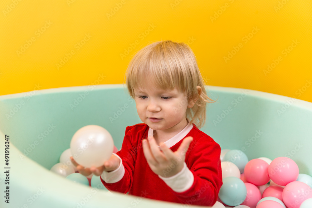 A little beautiful girl stands alone in dry pool with colorful balls and holds a white ball in her hands A little beautiful girl stands alone in dry pool with colorful balls and holds a white ball in her hands