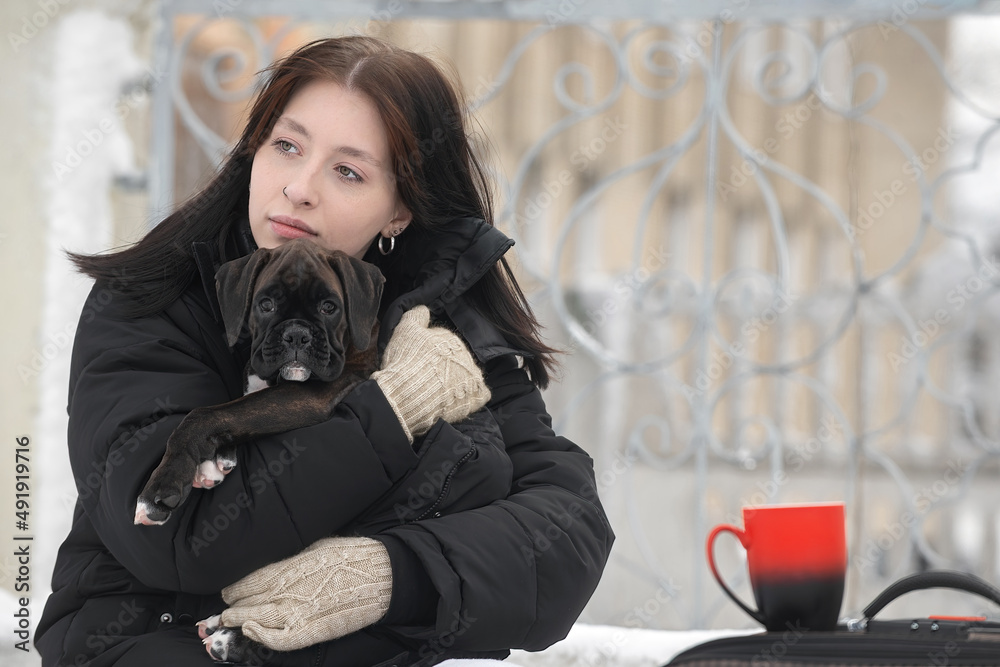 Φωτογραφία A beautiful young girl travels with her pet, a German boxer puppy, she hugged him and warms him, waiting for the train at the railway station, next to her is a suitcase and a red mug with a hot drink