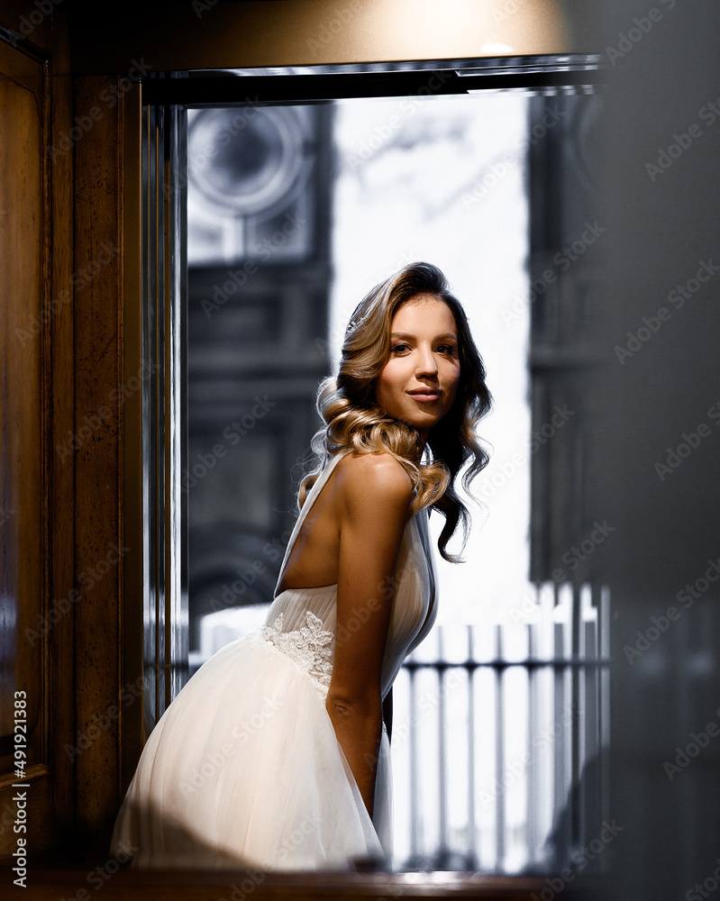 Side view of pretty and pensive bride, in ultra-sexy wedding dress with thin straps, which bending body forward , looking away while posing on background 