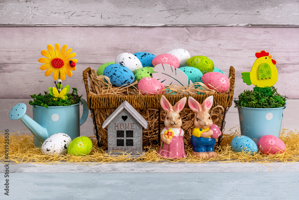 Fototapeta premium Easter eggs in a basket with decor. Colored eggs on straw. Decorative hares and house model. Beige wooden background.