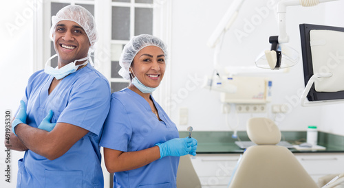 Two professional hispanic dentists in blue uniforms standing back to back in modern office of dental clinic