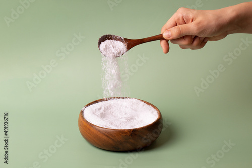 The girl with her hand pours white collagen powder with a wooden spoon into a wooden plate on a green background.