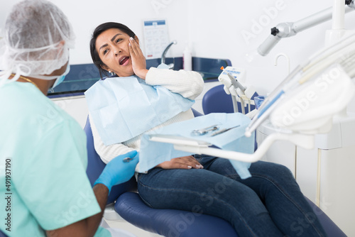 Upset hispanic woman sitting in dental chair  complaining to qualified dentist about toothache