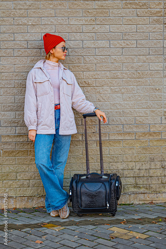 A young woman in casual clothes and with a suitcase poses at the airport at the train station against the background of a brick wall in sunny weather