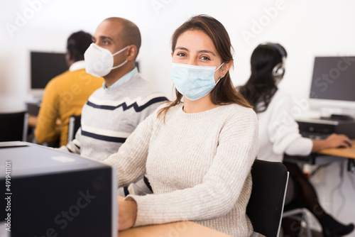 Positive female student wearing face mask working on computer in library. Concept of adult self-education during pandemic