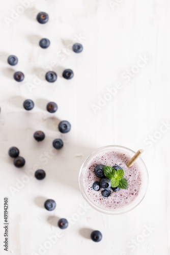 Glass of blueberry milkshake with fresh blueberries on white wooden background.
