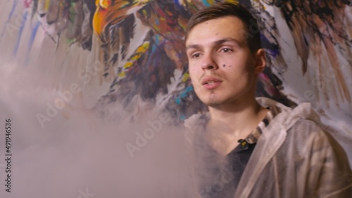 Artist designer draws an eagle on the wall. Craftsman decorator paints a picture with acrylic oil color. Vaper smokes vape. Painter painter dressed in a paint coat. Indoor. Dark magic cinematic look. © ivandanru