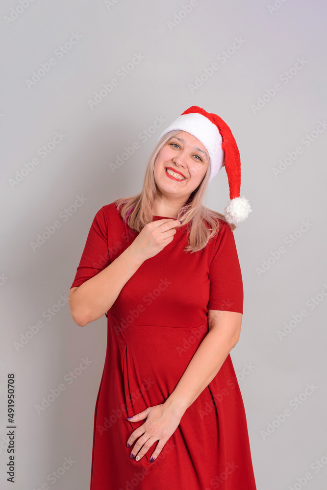 A beautiful adult woman in a Santa hat and a red dress poses on a gray background, vertical photo A beautiful adult woman in a Santa hat and a red dress poses on a gray background, vertical photo