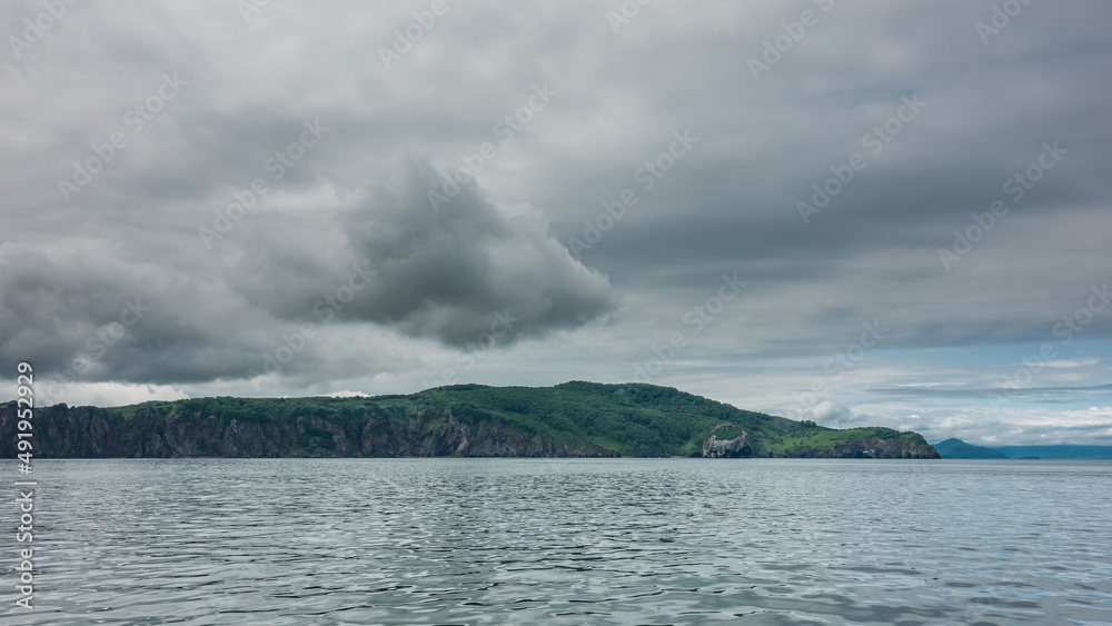 The hilly green coast of Kamchatka against a cloudy sky. Steep slopes of coastal cliffs. Ripples on the surface of the Pacific Ocean. Avacha Bay