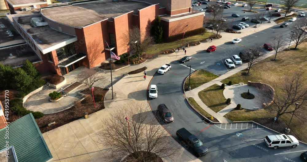 Carpool lineup outside school building in USA. American flag on pole during morning drop off. Education system in America.