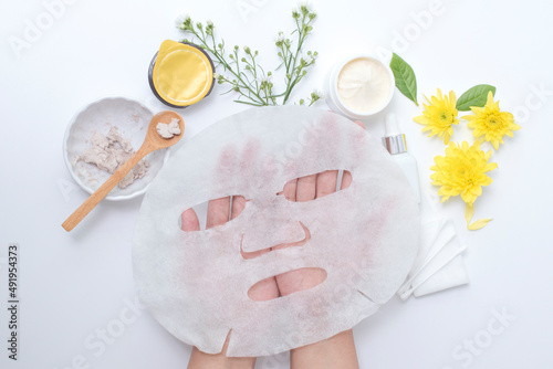 woman hand holding white sheet mask with clay mask wooden spoon yellow flowers and serum skincare cream products on white background. Top view