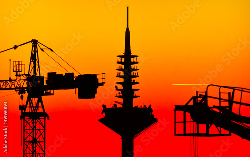 Die Silhouette der Kanzel und Spitze des Europaturms oder Ginnheimer Spargels in Frankfurt am Main, flankiert von den Silhouetten zweier Baukräne vor rot-orange verfärbtem Abend- oder Morgenhimmel photo