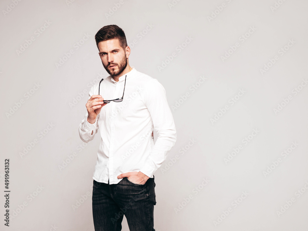 Portrait of handsome confident  model. Sexy stylish man dressed in white shirt and jeans. Fashion hipster male posing in studio on grey background. Holding sunglasses. Isolated