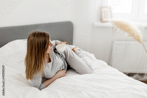 Young beautiful woman in light pajamas enjoys morning in bed with coffee in cozy bedroom. Morning rituals