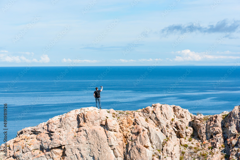 Man with backpack standing on a rock at the end of earth using his smartphone for selfie photoshoot. Hiking or travel concept. Exploring the great outdoors. Beauty of nature 