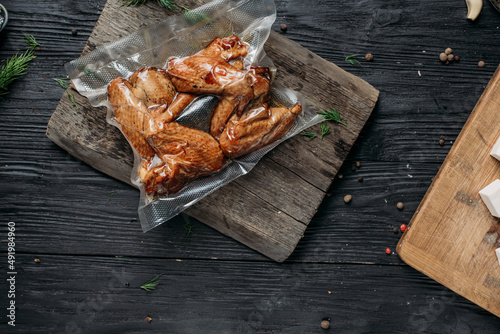 homemade hot-smoked chicken wings, in vacuum packaging on an old wooden board next to vegetables and spices