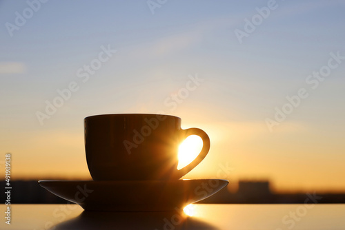 Silhouette of coffee or tea cup on background of sunset sky and shining sun. View from the window to evening city