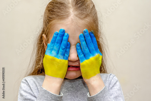 A child is crying with the flag of Ukraine on his hands (palms). The girl covered her face with her hands and cries. A tear. Children's tears of war. Children against war.