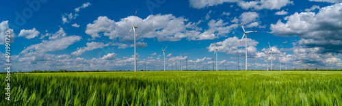 Panoramic view over beautiful farm landscape with green wheat field and wind turbines to produce green energy in Germany, Spring, blue dramatic sky and sunny day.