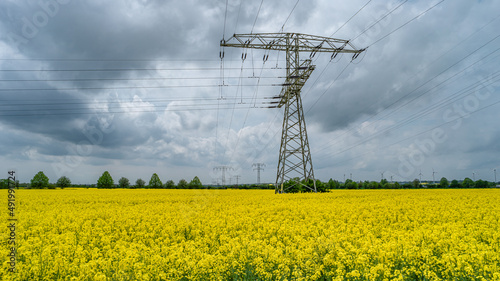 Beautiful farm landscape with yellow rapeseed at blossom field, wind turbines to produce green energy and high voltage power lines in Germany, at Spring and dramatic rainy sky.
