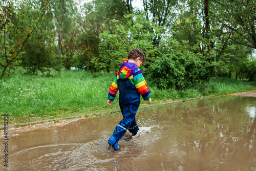 Fototapeta Naklejka Na Ścianę i Meble -  the boy runs and jumps through puddles in a waterproof suit and rubber boots.Bright kurta in the colors of the rainbow