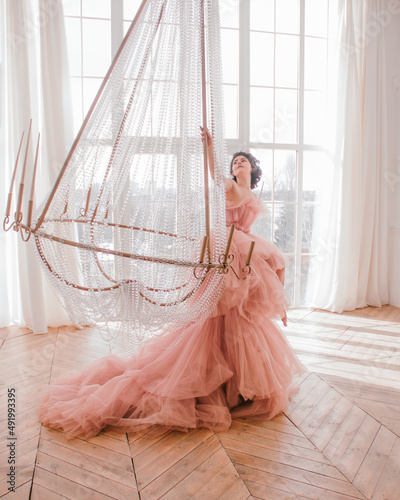 young woman with short black hair in pink princess dress with train is sitting on the crystal chandelier near huge panoramic window background like dior style. fashion concept, free space