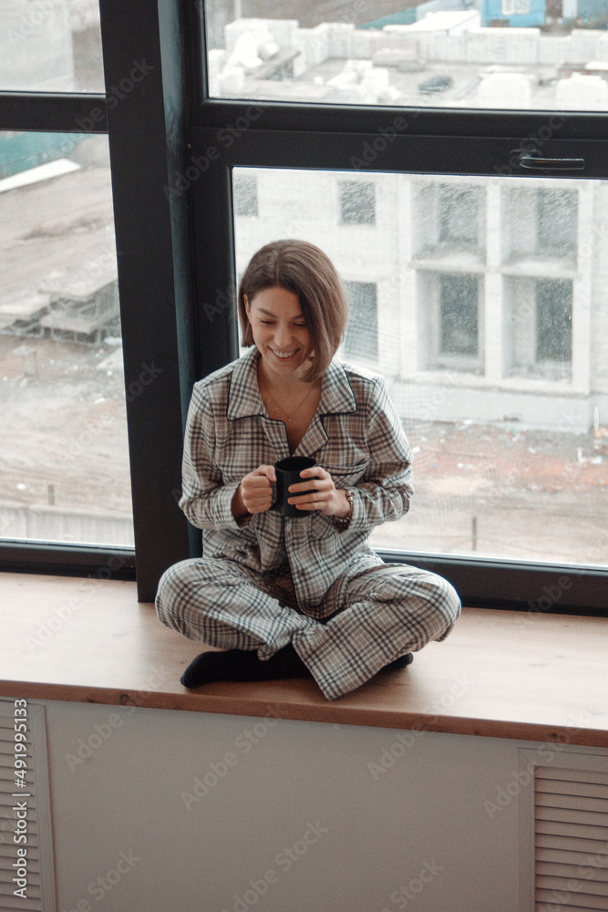 young curly brunette girl in gray plaid pajamas is sitting with cup in hands on the huge window background and smiling happy, camera from top. lifestyle concept, free space