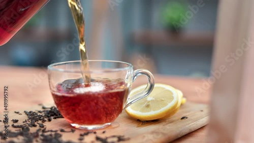 Closeup pouring fresh hot aroma black tea from jar teapot into glass cup on wooden table slowmo photo