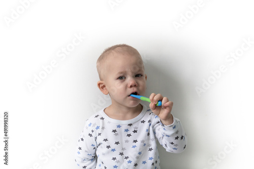 A little boy stands against a white wall, dressed in a T-shirt with stars, brushes his teeth and takes care of his mouth.