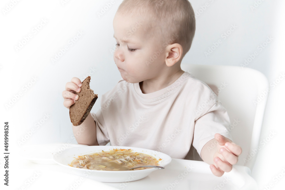 A little boy eats soup with bread on his own in a highchair against the background of a white wall. A little boy eats soup with bread on his own in a highchair against the background of a white wall.