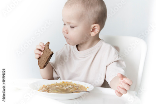 A little boy eats soup with bread on his own in a highchair against the background of a white wall.