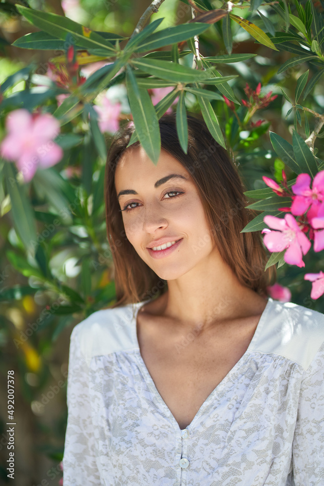 Pretty young woman with perfect skin and charming smile posing near blooming pink flowers in garden. Nude make up Pretty young woman with perfect skin and charming smile posing near blooming pink flowers in garden. Nude make up