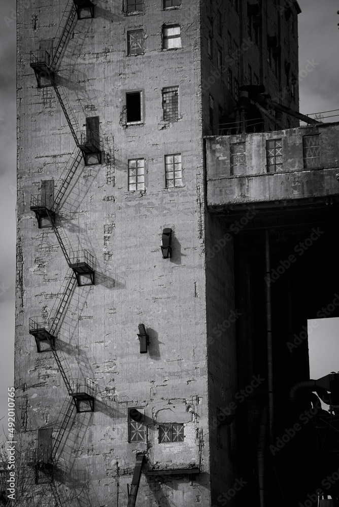 Black and white street photo with the charm of the ugly architecture of old factory buildings. The concrete structure of the plant with a metal staircase and different windows. Conceptual photo with a