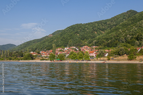Picturesque view on village on shore Danube river. Green forest and hills. Summer day. Travel and holiday. Serbia.