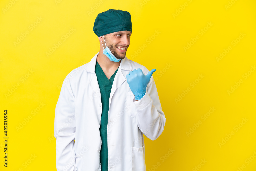 Surgeon blonde man in green uniform isolated on yellow background pointing to the side to present a product Surgeon blonde man in green uniform isolated on yellow background pointing to the side to present a product