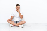 Young blonde man sitting on the floor isolated on white background having doubts and with confuse face expression
